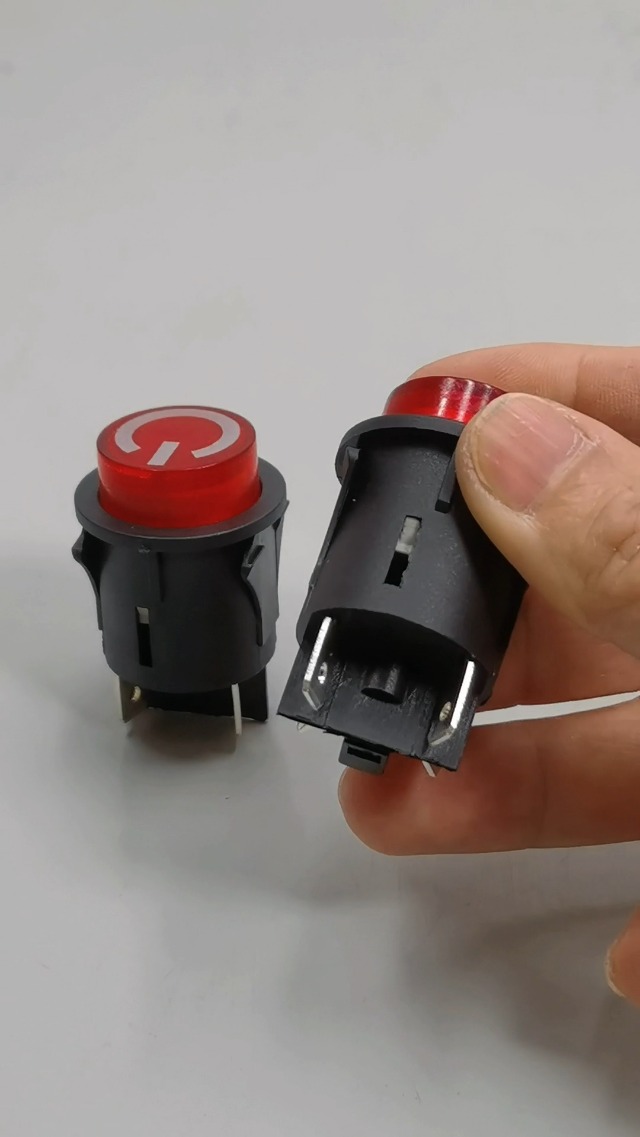 A hand holding a SHENGLE Power Button Start Switch, showing the red button and the black housing with metal terminals on the back.