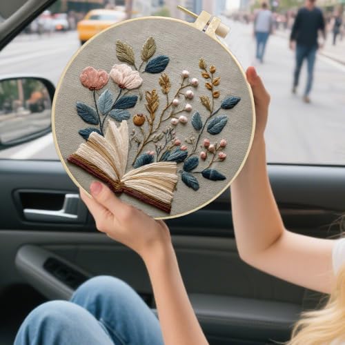 Person holding a round embroidery hoop with raised flowers and an open-book design while seated inside a car.