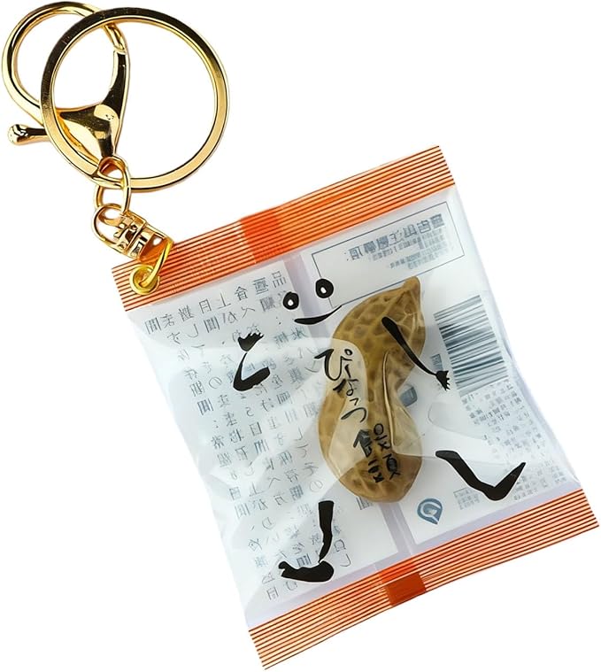Gold keychain with a split ring and clasp attached to a small clear snack-style wrapper containing a realistic peanut, with cartoon arms and legs and Japanese text printed on the wrapper.