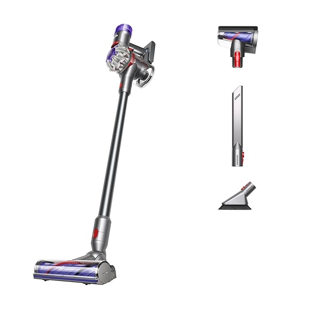 Dyson V8 Plus cordless stick vacuum in silver and yellow finish on a US bedroom carpet, the detangling motorbar cleaner head pulled forward across medium-pile pet hair, the detachable handheld unit released and held in one hand to clean a couch cushion in the background, the user-replaceable battery visible in the slot below the trigger — Dyson's most-reviewed cordless stick in this guide with 5,031 verified Amazon reviews, refreshed with the detachable battery that addresses the original V8's sealed-battery reliability complaint.