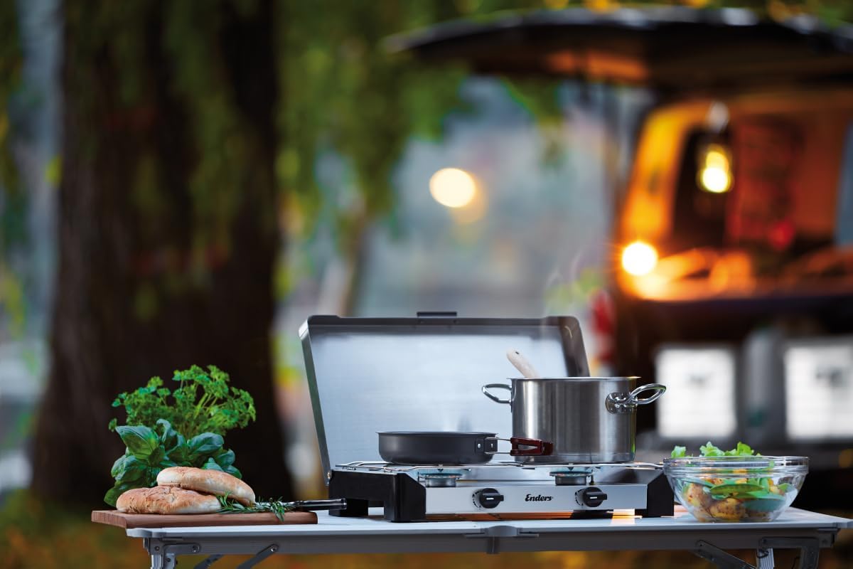 The Enders Brisbane camping stove in an outdoor setting, with two pots on the burners, surrounded by fresh vegetables and bread, suggesting a meal being prepared.