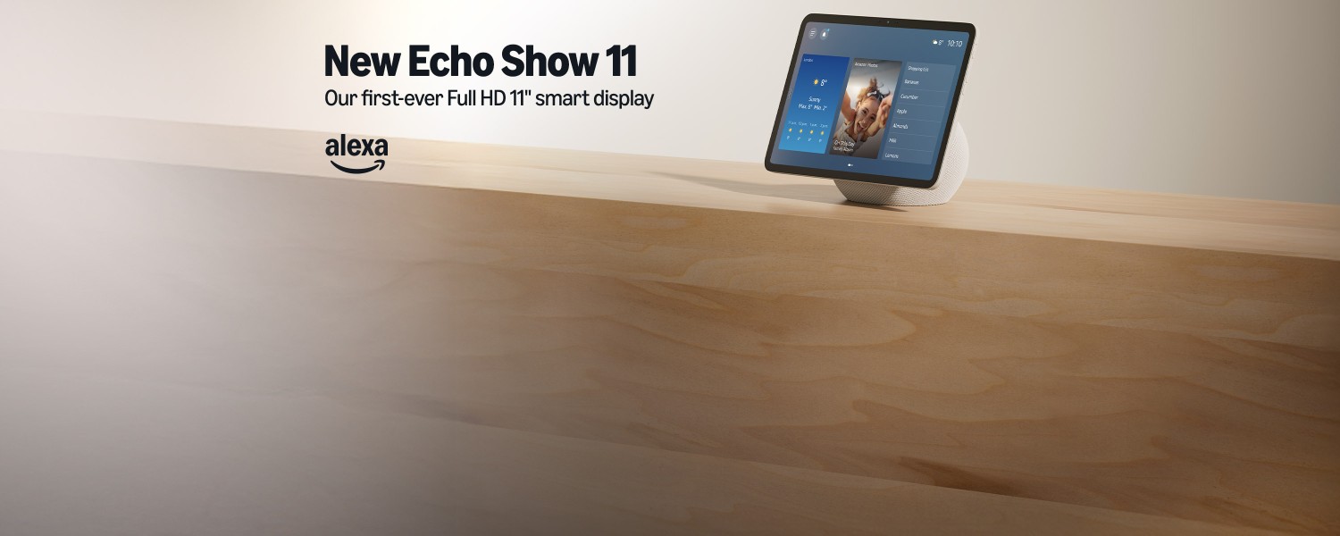 Echo Show 11 smart display on a wooden shelf. The device features a white base with a large 11-inch Full HD touchscreen. Text reads: "New Echo Show 11" and "Our first-ever Full HD 11" smart display".