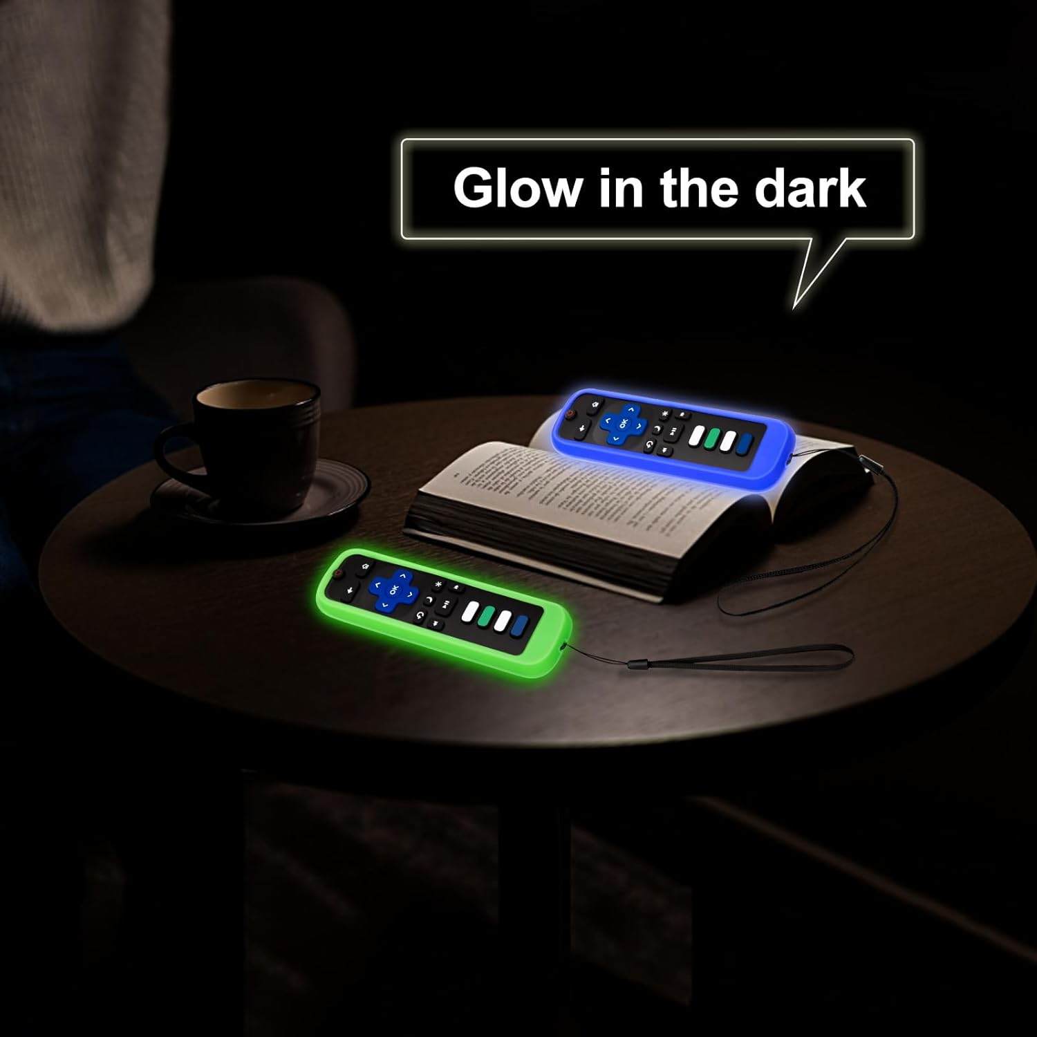 Two Roku remotes with green and blue silicone covers glowing brightly in a dark room, placed on a table next to a book and coffee cup.