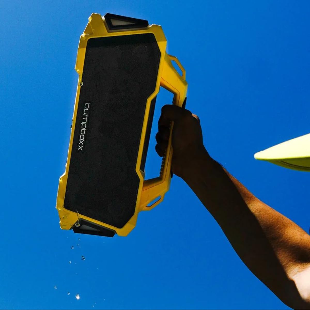 Hand holding a yellow Bumpboxx Thump speaker with water dripping, demonstrating water resistance