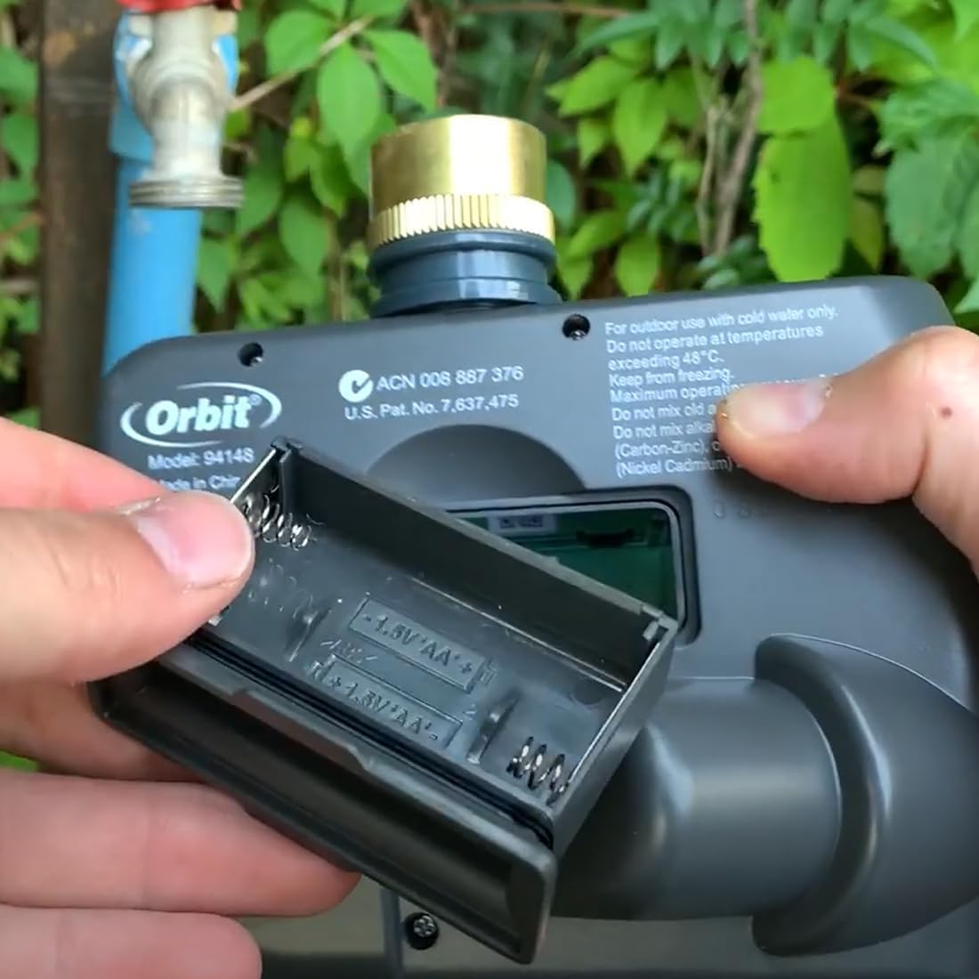 Hands opening the battery compartment of the GNCGarden Orbit Buddy II irrigation programmer to insert AA batteries.
