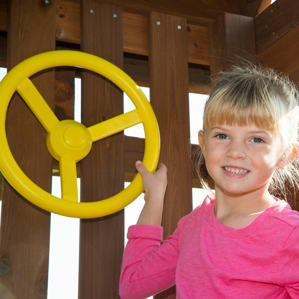 A young girl smiling while holding a yellow toy steering wheel on the playset.