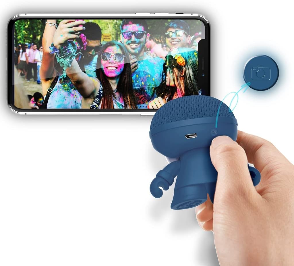 A hand holding the Xoopar Mini Xboy Eco speaker, with a blue button highlighted, next to a smartphone displaying a group photo, indicating the speaker's selfie remote function.
