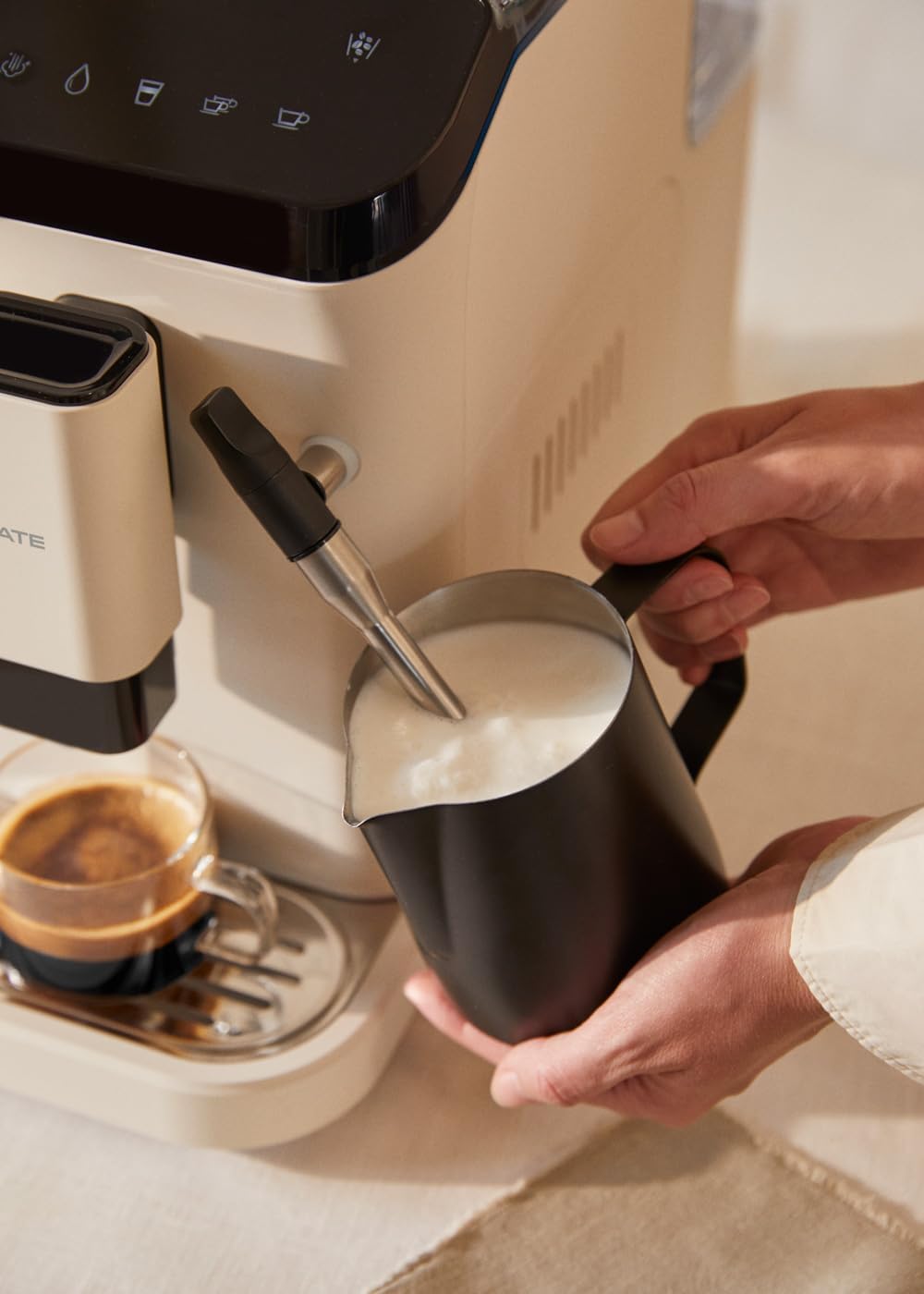 A person using the steam wand of the CREATE THERA MATIC TOUCH coffee machine to froth milk in a black pitcher.