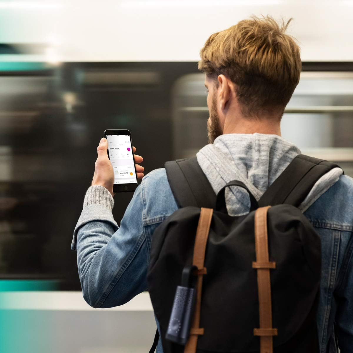 A person with a backpack, holding a smartphone and looking at the screen, with the Plume Labs Flow 2 sensor clipped to their backpack, indicating portable use.