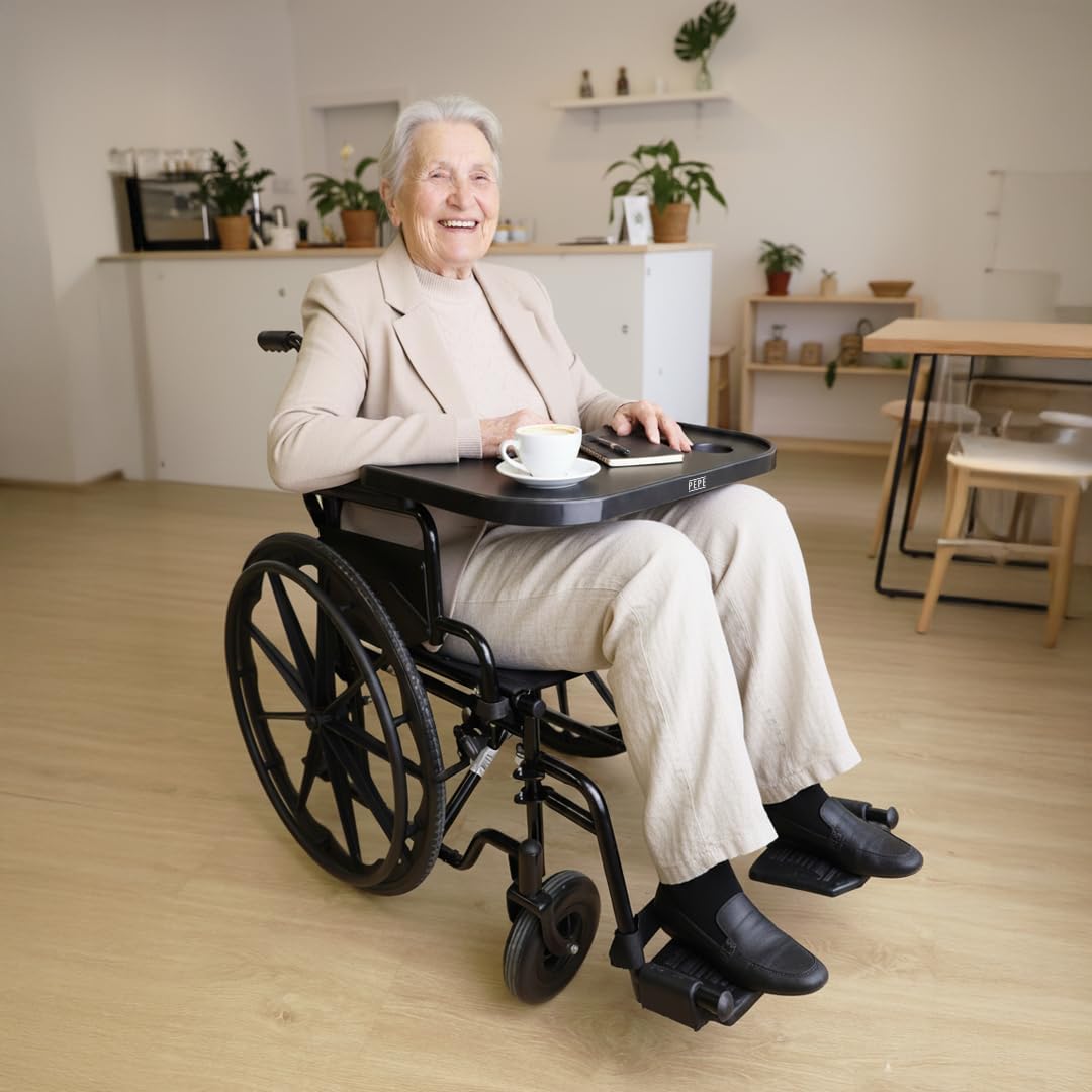 An elderly woman smiling while sitting in a wheelchair with the tray in use, holding a cup of coffee.