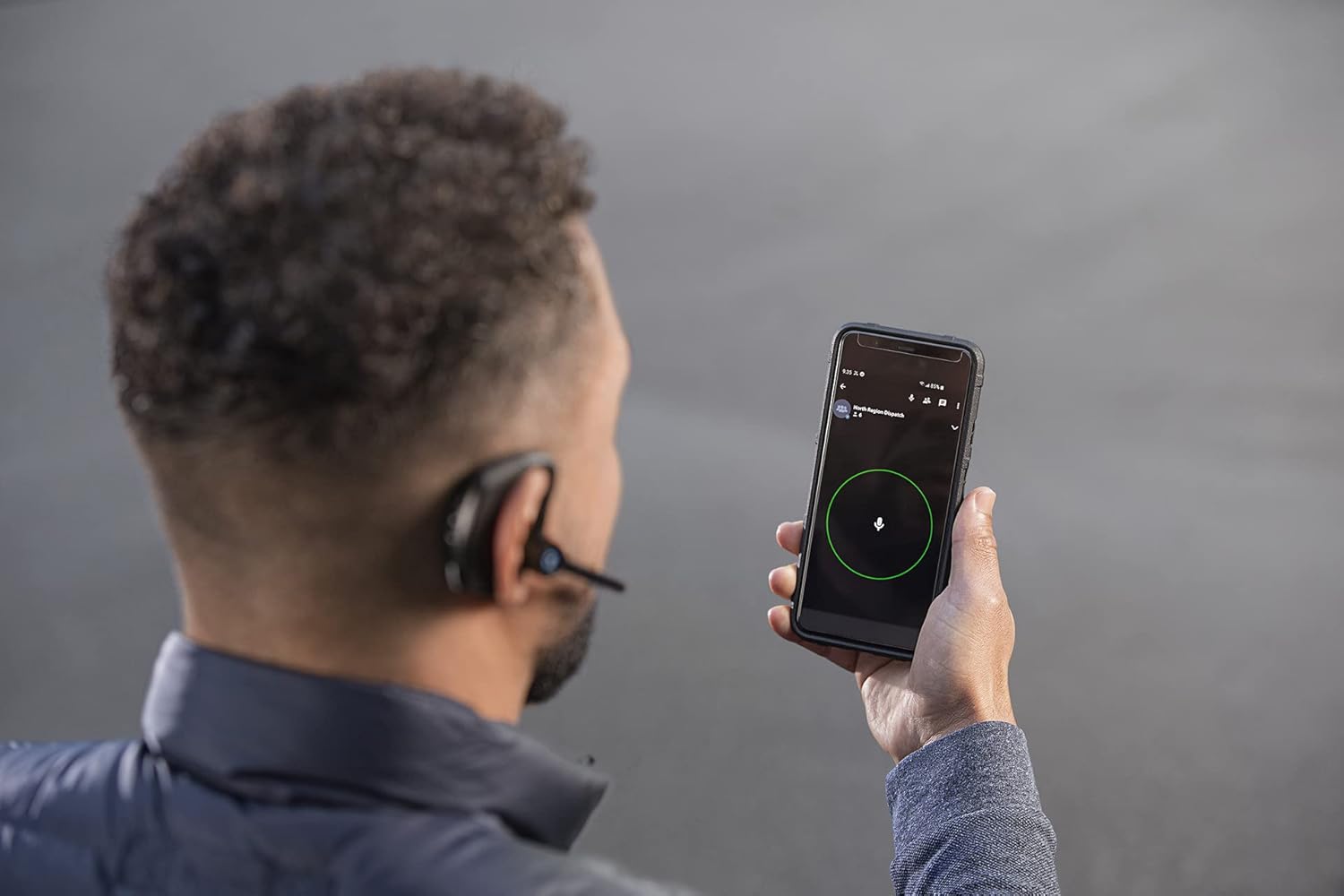 A person holding a smartphone with the BlueParrott M300-XT headset visible in the background, indicating app connectivity.