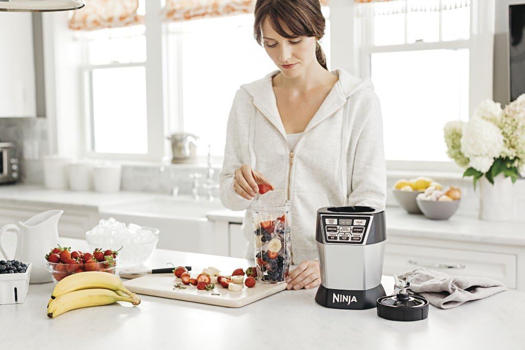 A woman adding strawberries to a Nutri Ninja cup on a kitchen counter, with the Ninja Nutri Bowl Duo motor base nearby.