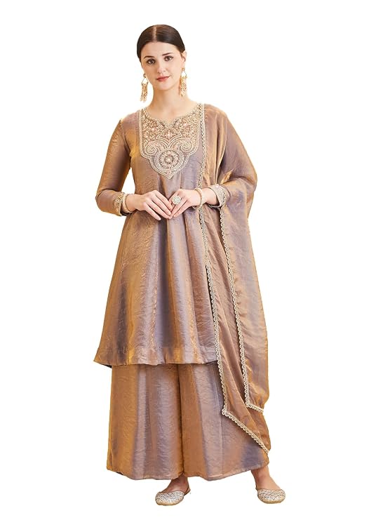 Woman wearing a golden-beige silk kurta, sharara, and dupatta set. The kurta features intricate embroidery at the neckline, three-quarter sleeves, and a flared silhouette. The dupatta has a gold lace border.