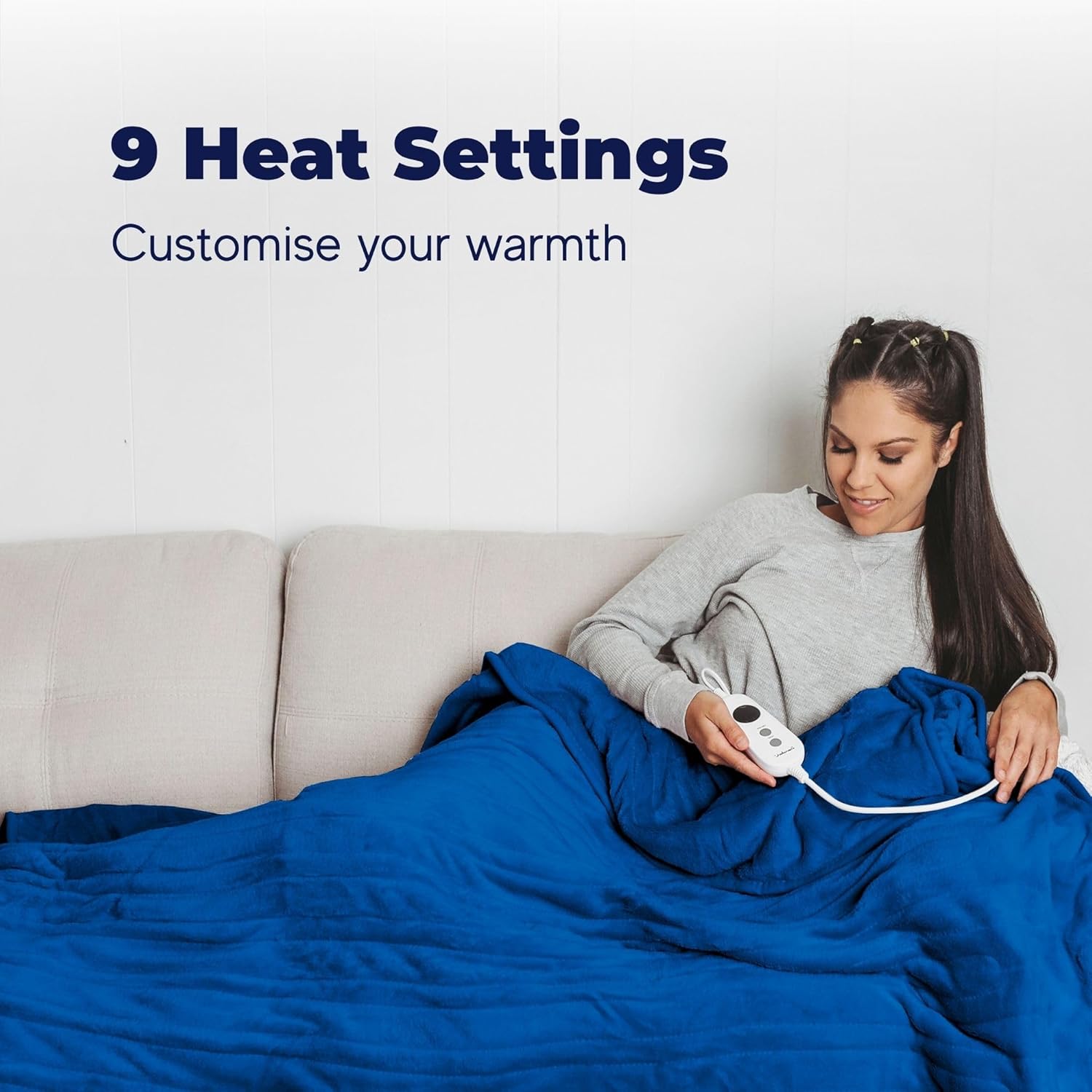 A woman sitting comfortably on a couch, covered by a blue Dreamaker heated blanket, holding the white controller in her hand.