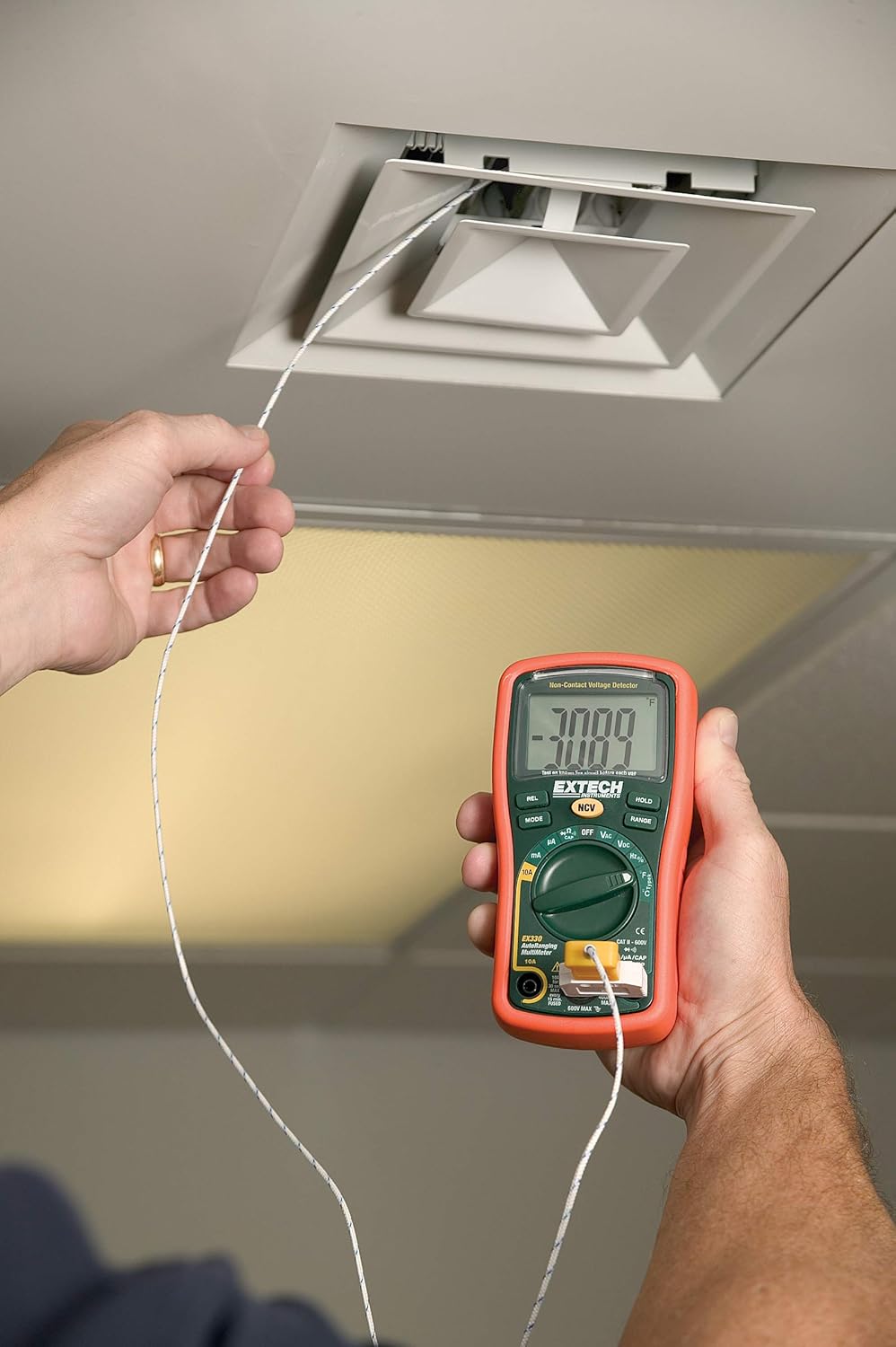A person holding the Extech EX330 Multimeter with a Type K temperature probe inserted into an air vent, measuring the temperature. The display shows '70 F'.