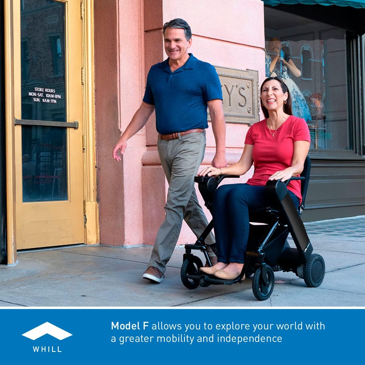 A woman in the WHILL Model F Power Chair and a man walking beside her on a city sidewalk, demonstrating ease of movement.