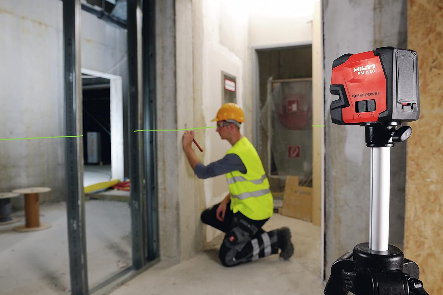 Hilti PM 2 Green Line Laser Level mounted on a tripod, projecting a horizontal green line across a room where a worker is marking a wall.