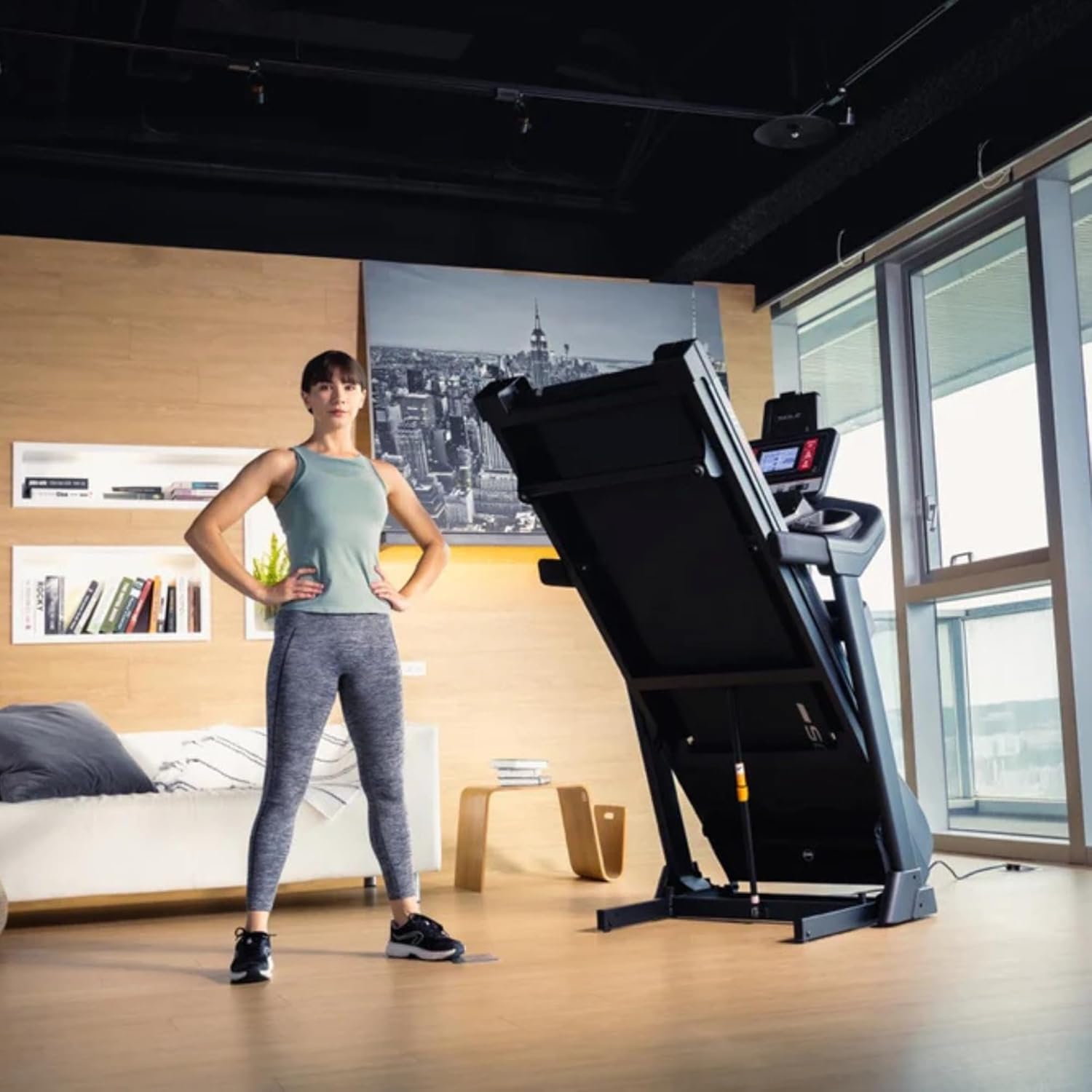 A person standing in a modern home gym beside a folded Sole F63 Treadmill, showcasing its space-saving design, with wooden flooring, large windows, a cityscape wall art, and a cozy seating area in the background.
