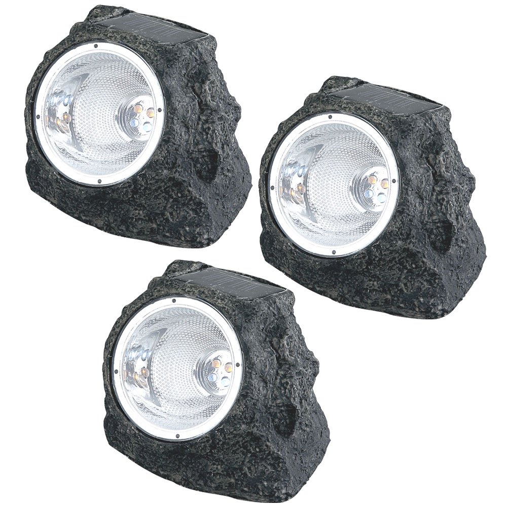 Three Globo Solar stone spotlights placed in a garden setting among green plants and small rocks.