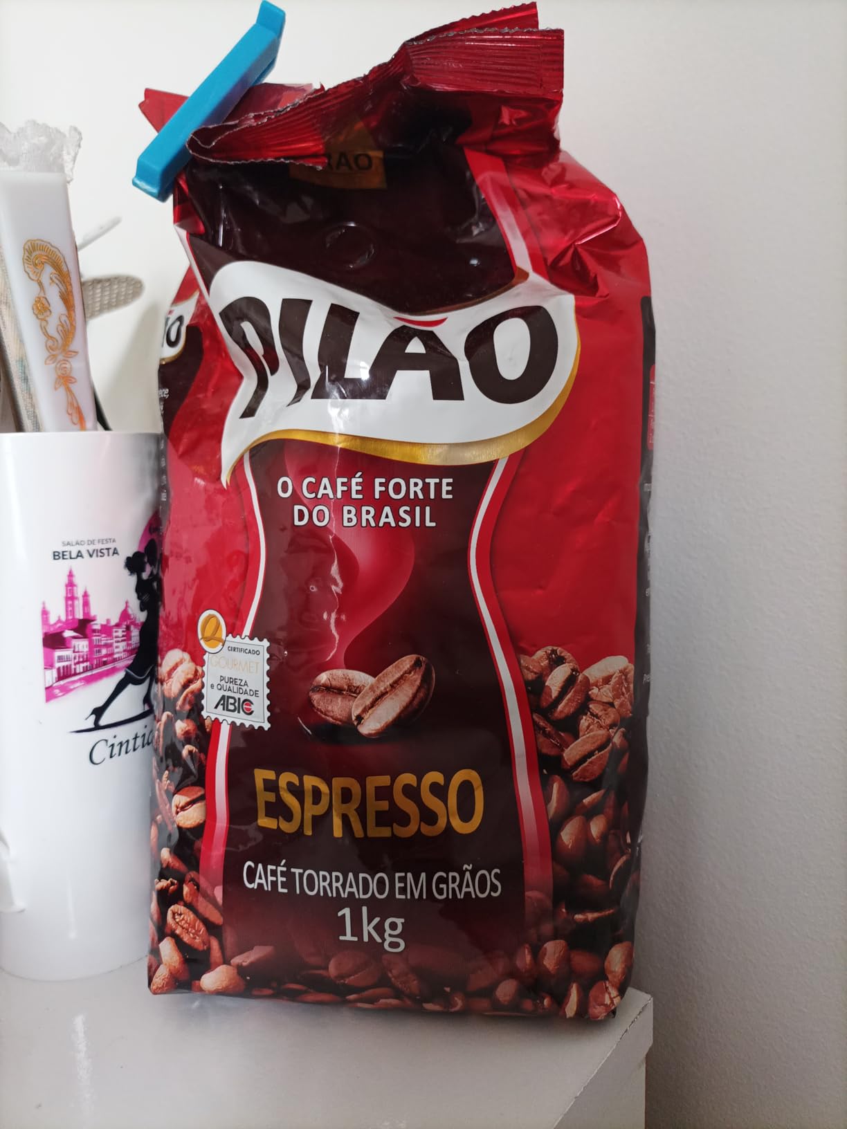 Pacote de 1kg de café em grãos Pilão Espresso posicionado sobre uma superfície ao lado de um copo personalizado