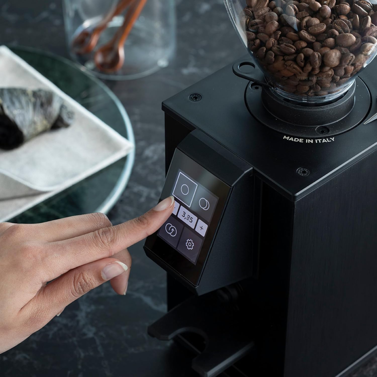 A hand touching the screen of the MACAP LEO 55 Touch coffee grinder, showing the digital display with dose settings and a timer of 3.85 seconds.