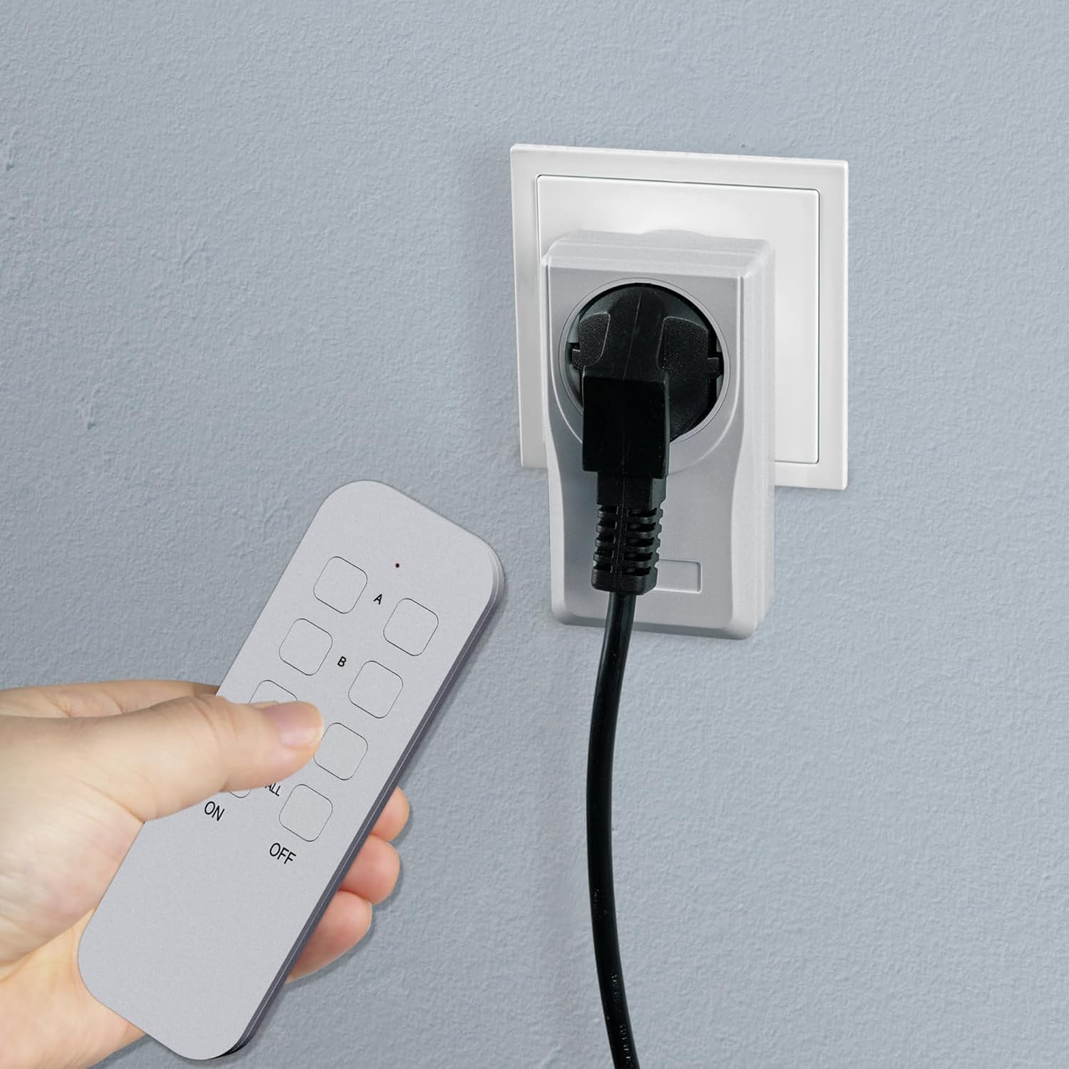 A hand holding the remote control, pressing a button, with a remote control outlet plugged into a wall socket in the background.