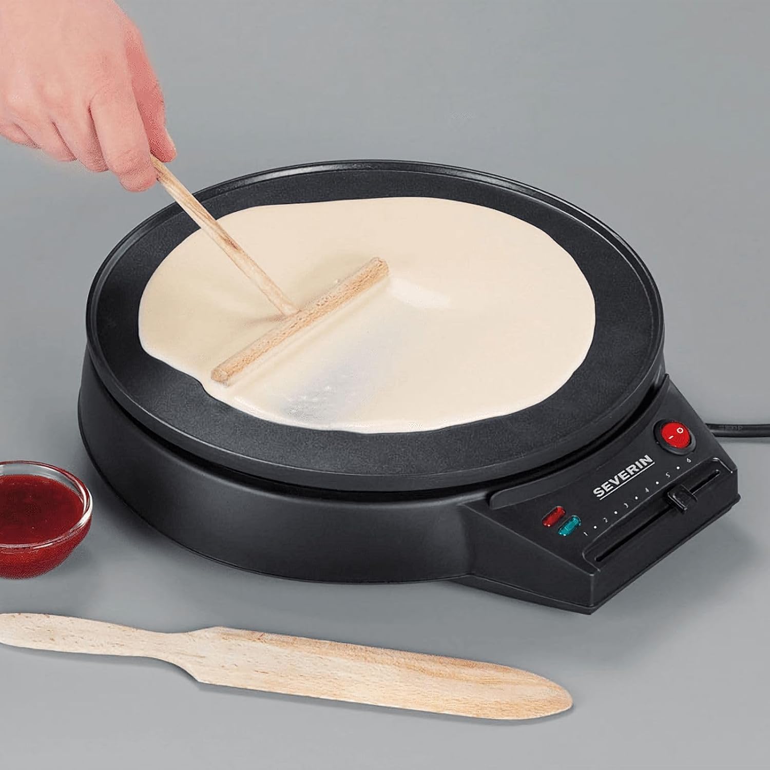 A hand using the wooden turning stick to spread crepe batter on the hot surface of the crepe maker.
