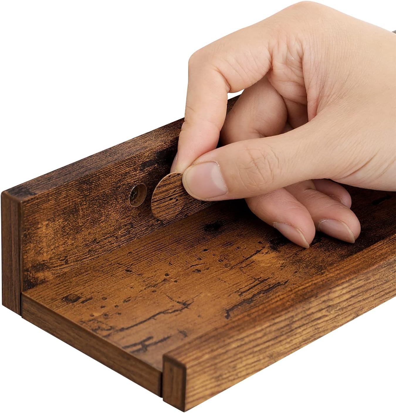 A hand placing a decorative cap over a screw hole on the rustic brown floating shelf, demonstrating how to conceal the mounting hardware.