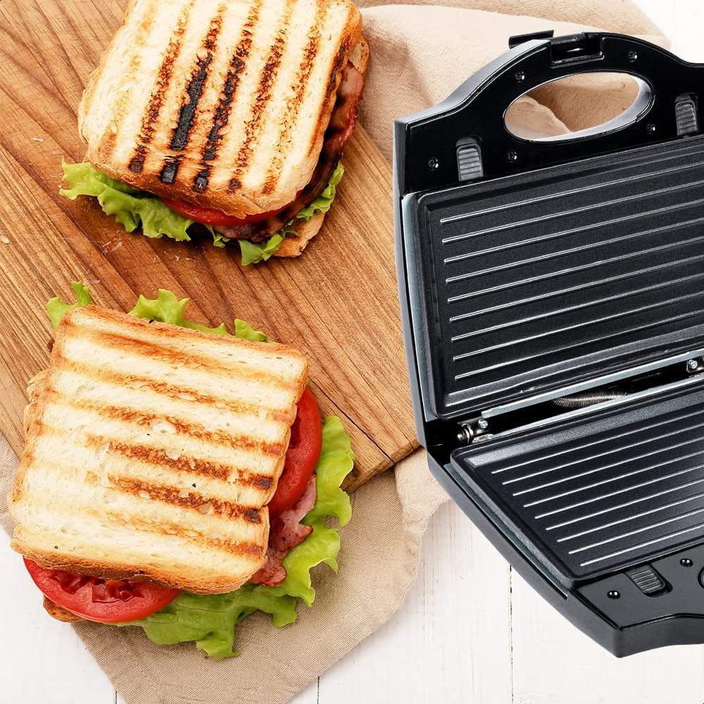 Two grilled sandwiches with lettuce and tomato on a wooden board next to the open sandwich maker.