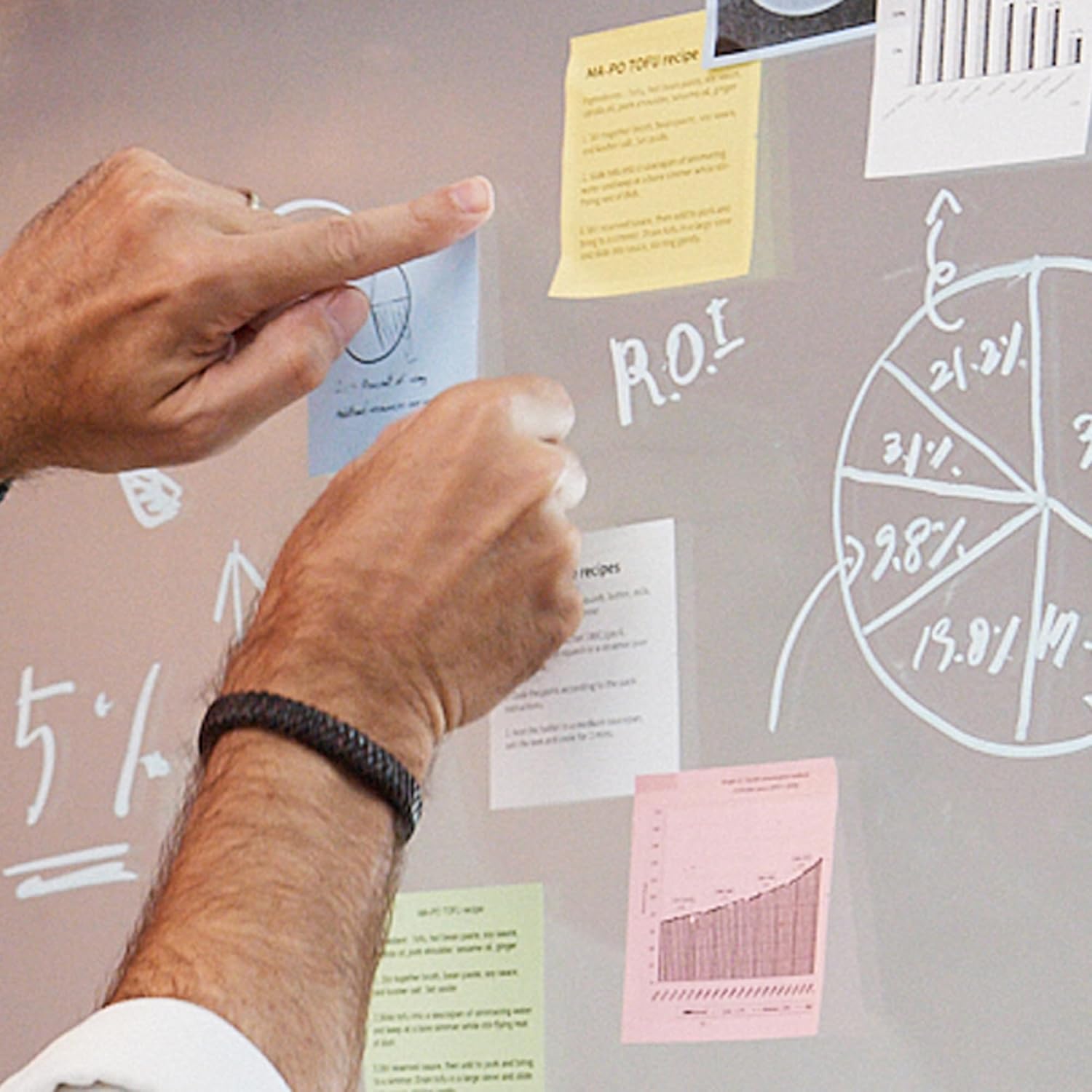 Two pairs of hands interacting with sticky notes and diagrams on a clear board.