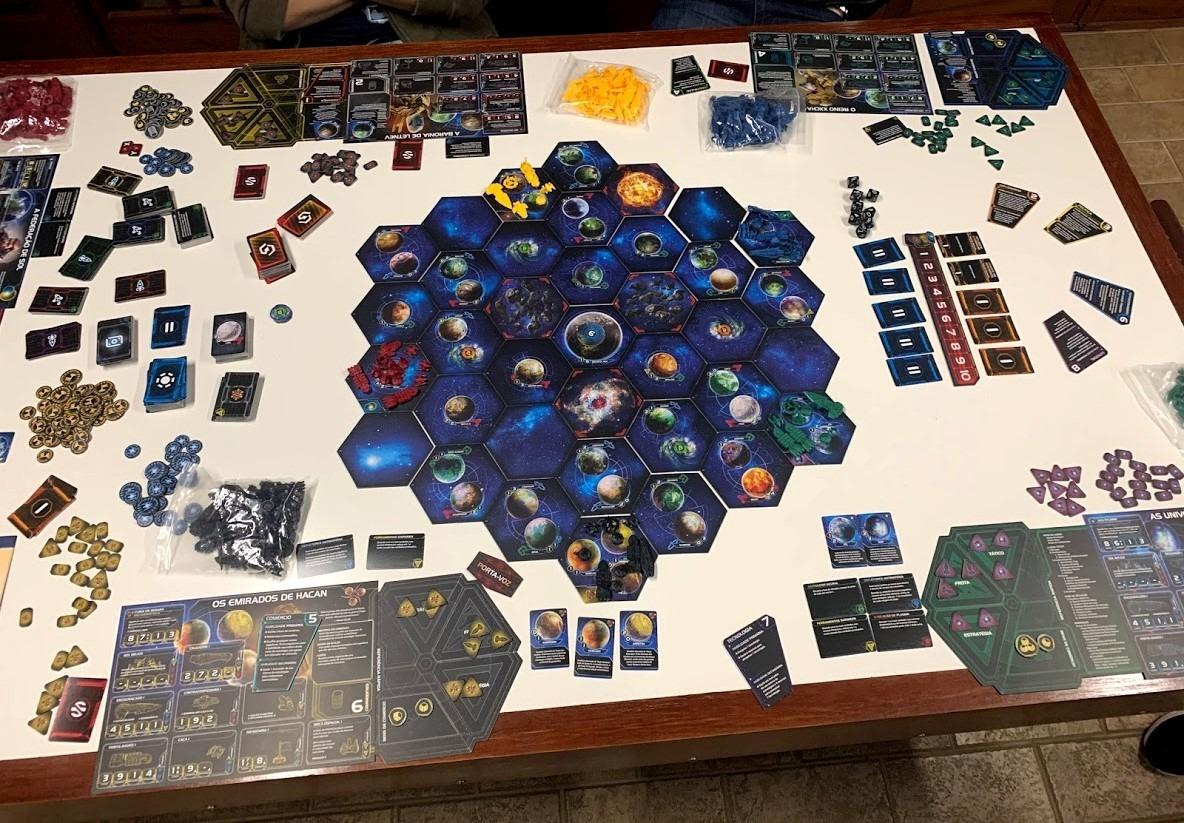 Partida em andamento do jogo Twilight Imperium 4ª Edição organizado sobre uma mesa branca, exibindo o mapa galáctico hexagonal, cartas e componentes.