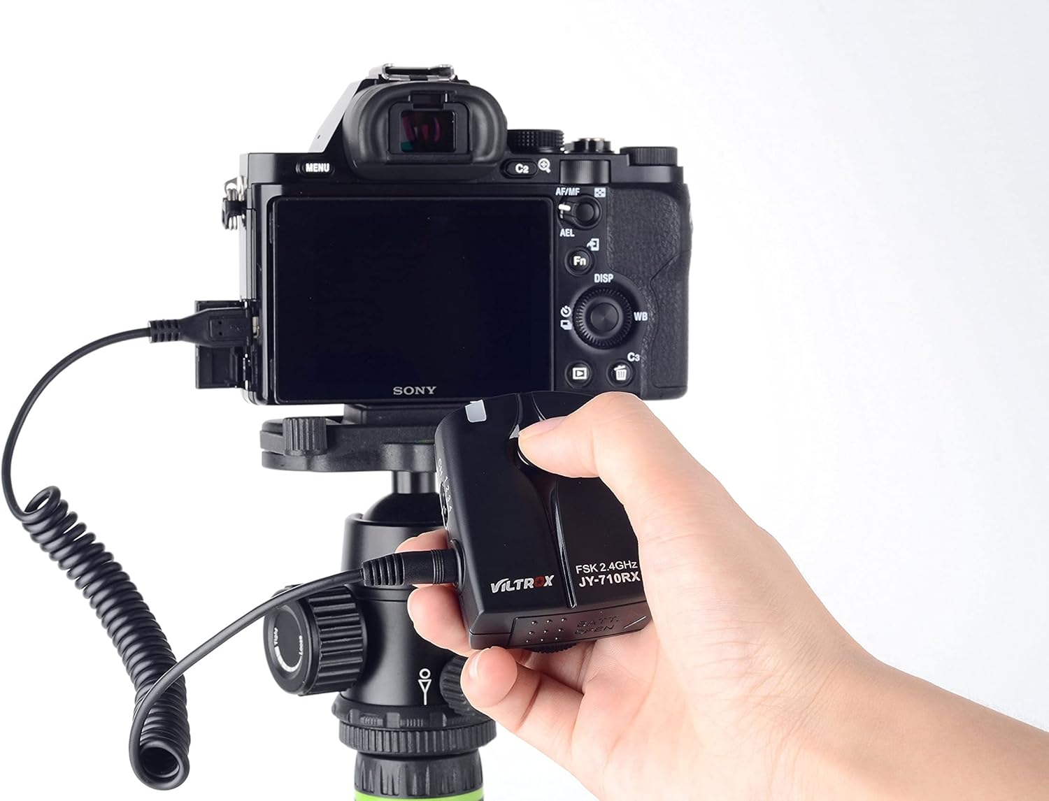A hand holding the VILTROX JY-710-S2 transmitter, which is connected via a coiled cable to a Sony camera mounted on a tripod.