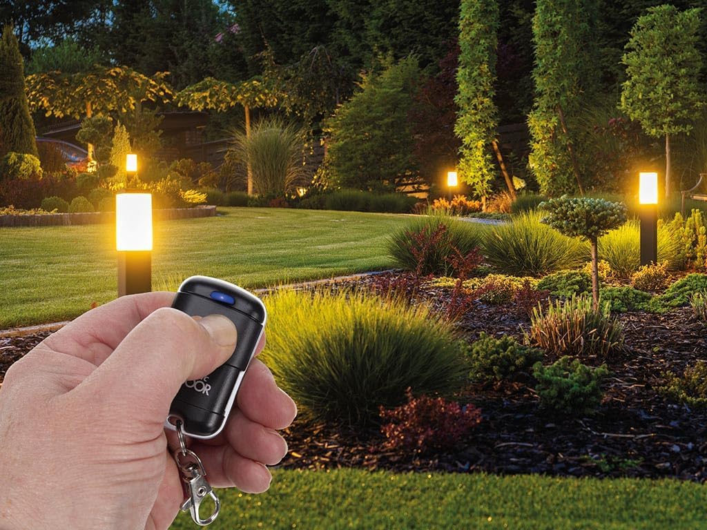 A hand holding the Unitronic remote control in a landscaped garden with illuminated path lights, showing its application for outdoor lighting.