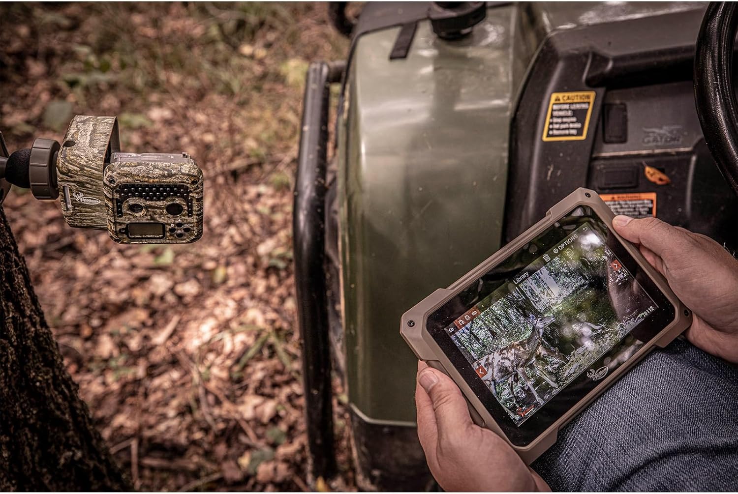 Wildgame Innovations Hex Trail Camera mounted on a tree, with a person viewing images on a tablet nearby.