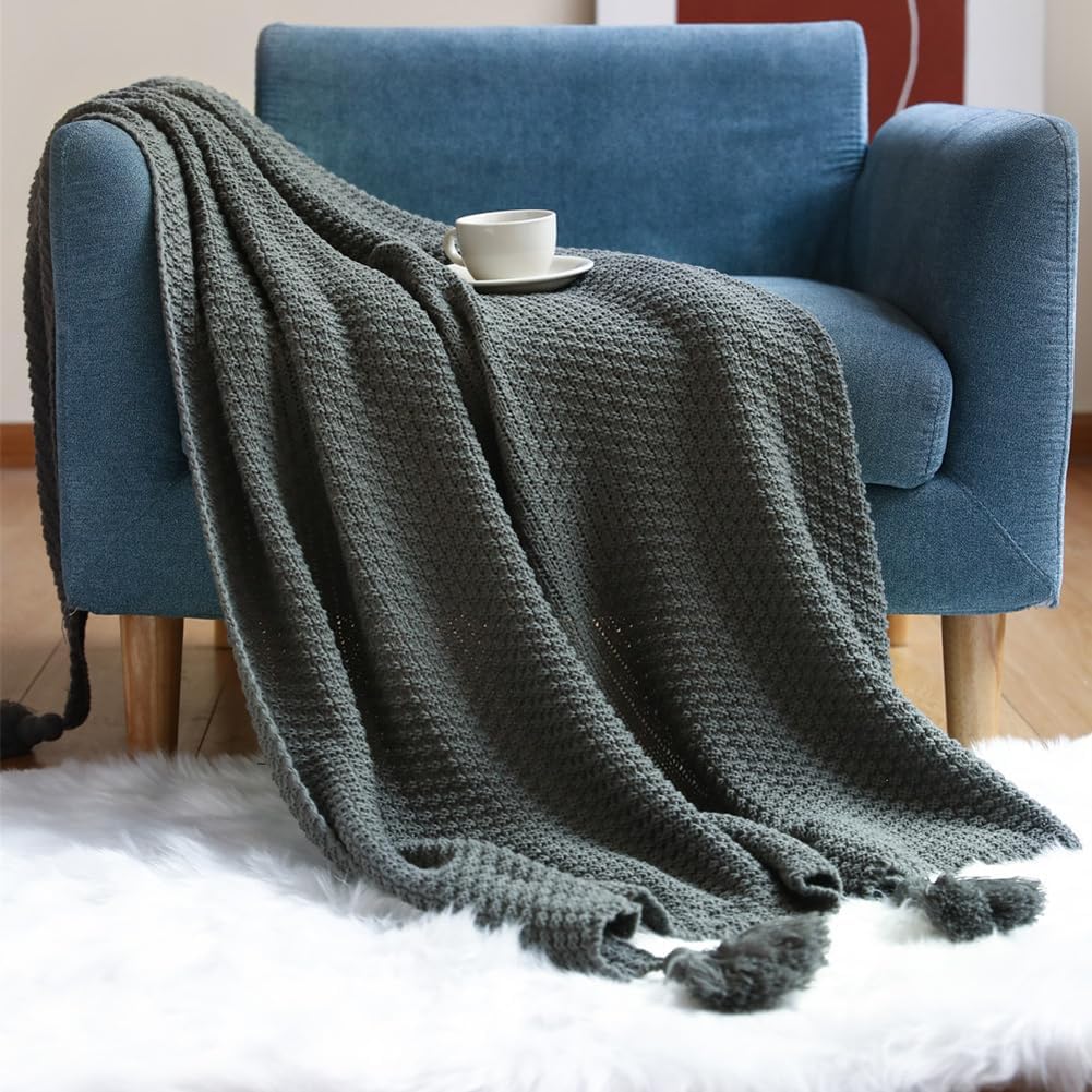 Dark grey Boho throw blanket draped over a blue armchair, with a white rug underneath and a white mug on a saucer on the blanket.
