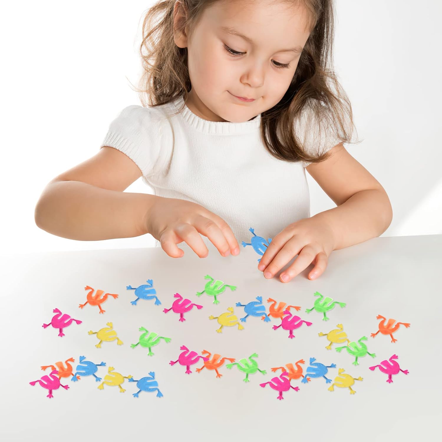 A young girl playing with multiple colorful jumping frogs on a white table, demonstrating active engagement with the toys.