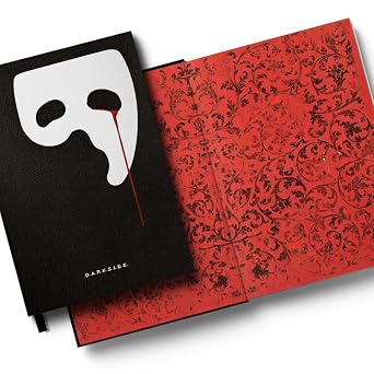 Livro "O Fantasma da Ópera" aberto, mostrando a capa preta com a máscara e uma gota de sangue, e a página interna com um padrão floral vermelho e preto.