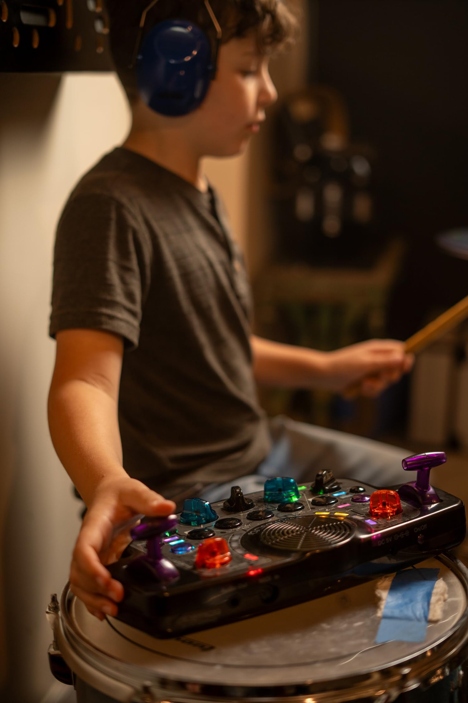 A young boy wearing headphones, playing drums, with the Blipblox After Dark Synthesizer placed on a drum.