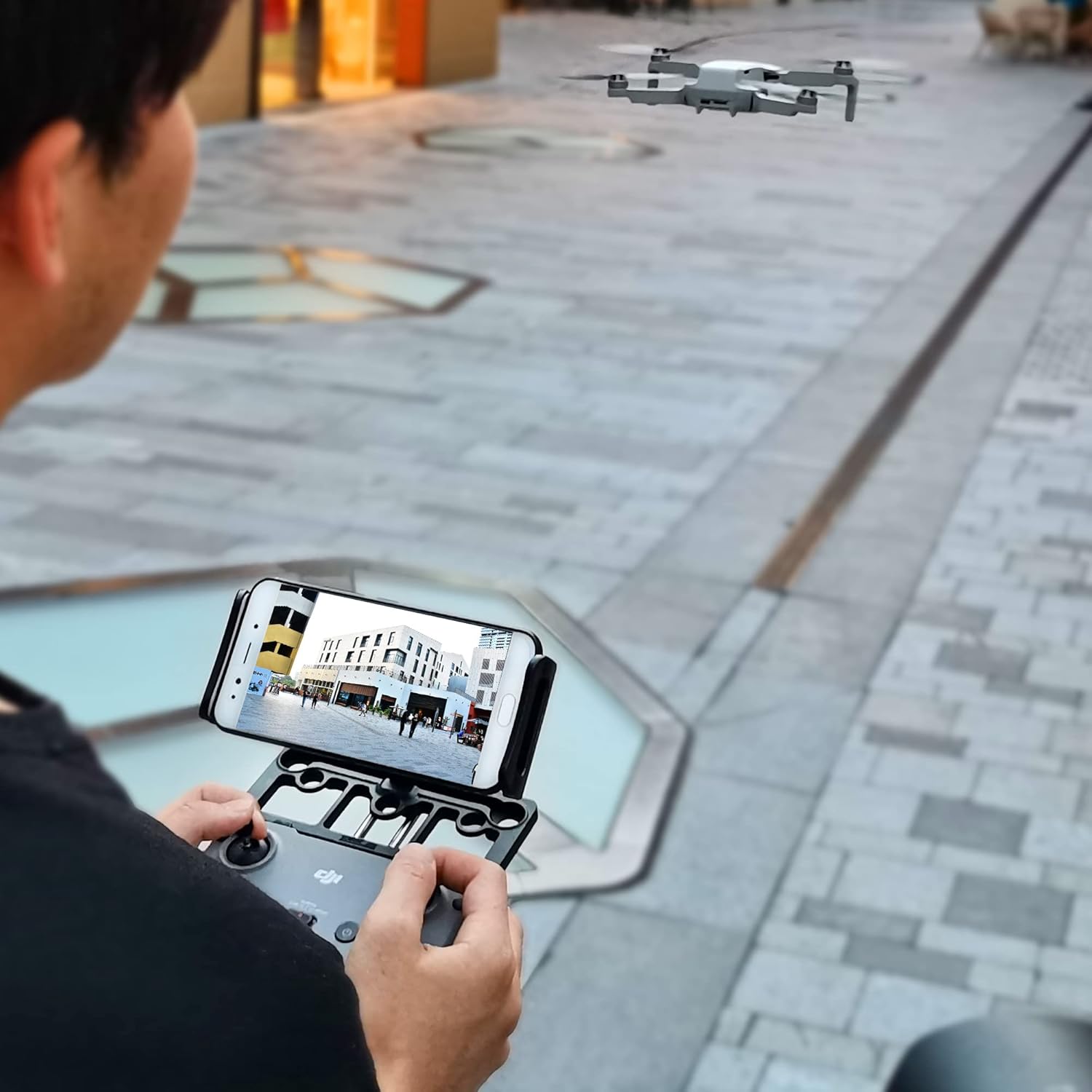 A person operating a DJI drone remote controller with the STARTRC Tablet Stand holding a smartphone, while a drone flies in the background.