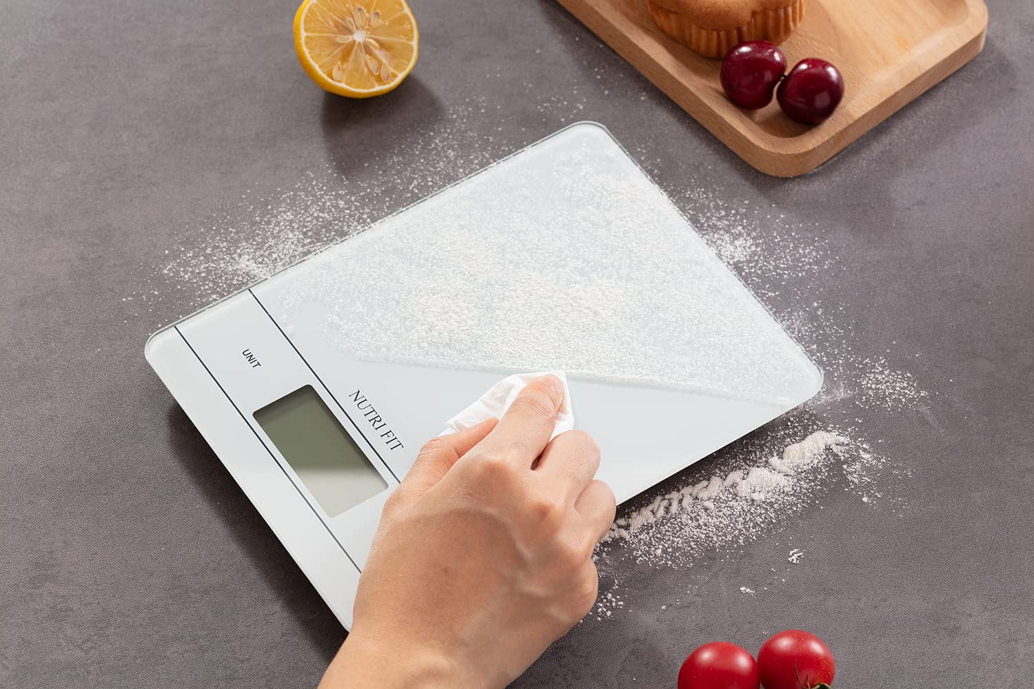 A hand wiping flour off the white tempered glass surface of the NUTRI FIT kitchen scale with a white cloth.