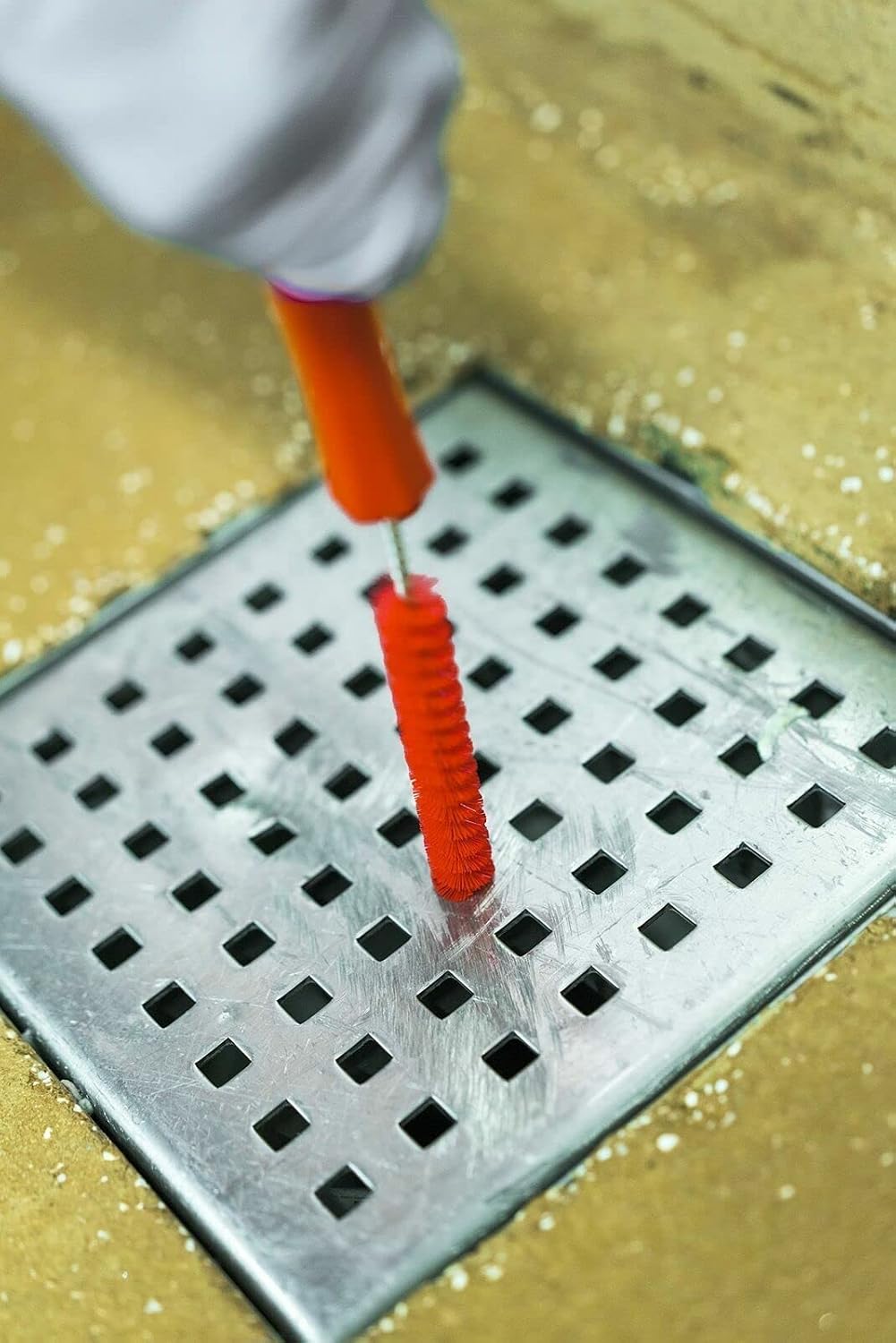 A person uses a red Vikan tube brush to clean a metal drain grate on a tiled floor.