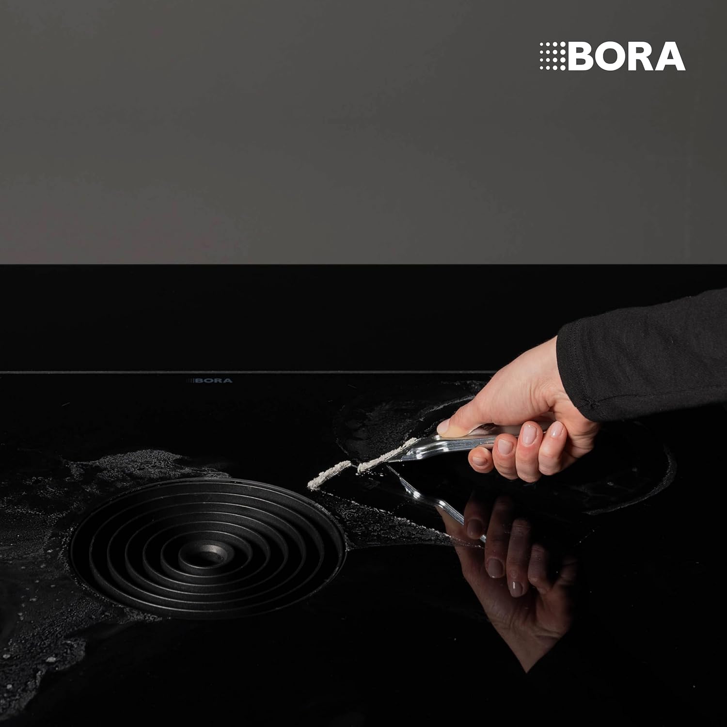 Hand using the BORA scraper to remove residue from a black cooktop.