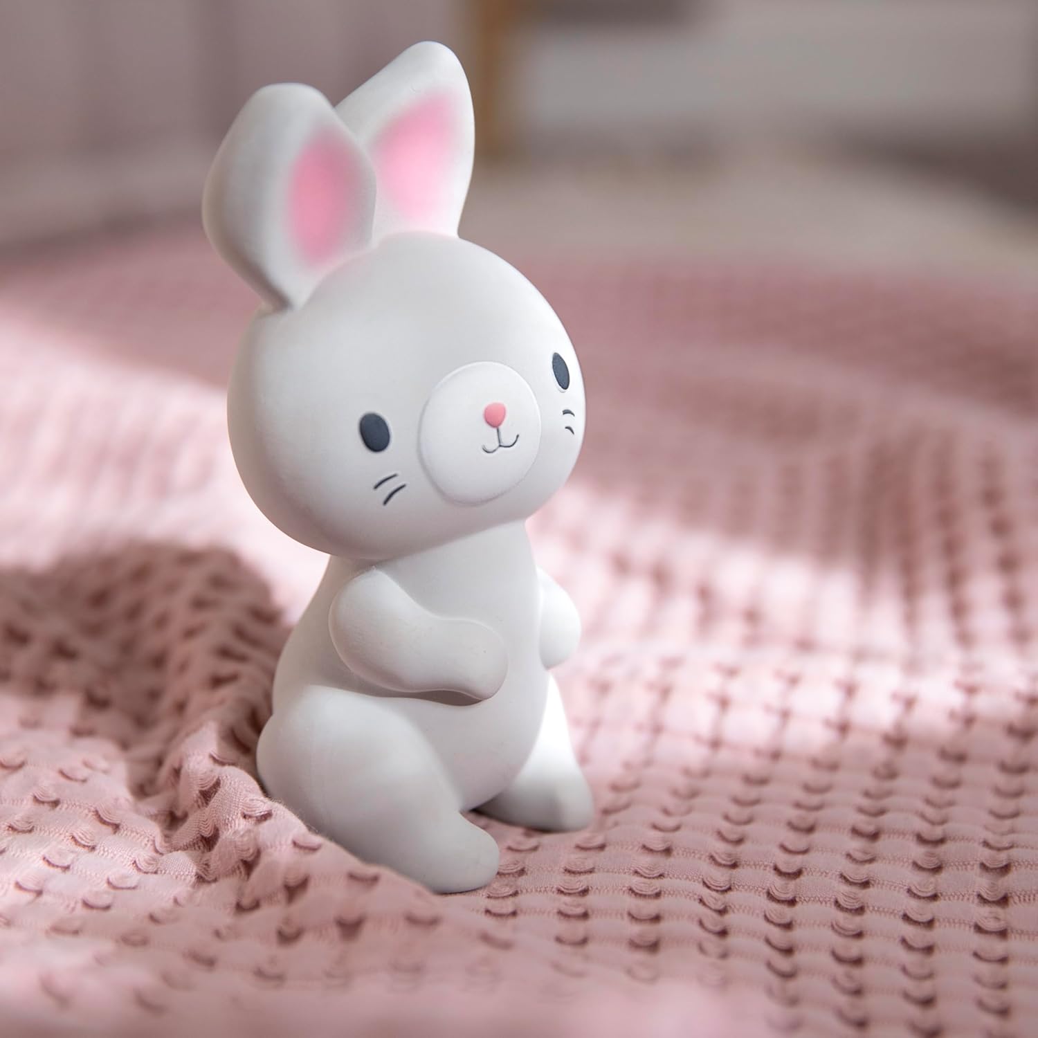 The Ingenuity Sylvi teether, a gray rabbit with pink ears and nose, standing upright on a pink knitted blanket.