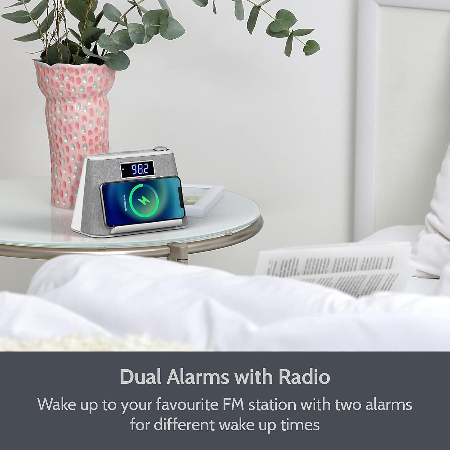 i-box alarm clock radio on a bedside table, displaying an FM radio frequency, with a smartphone on its charging stand.