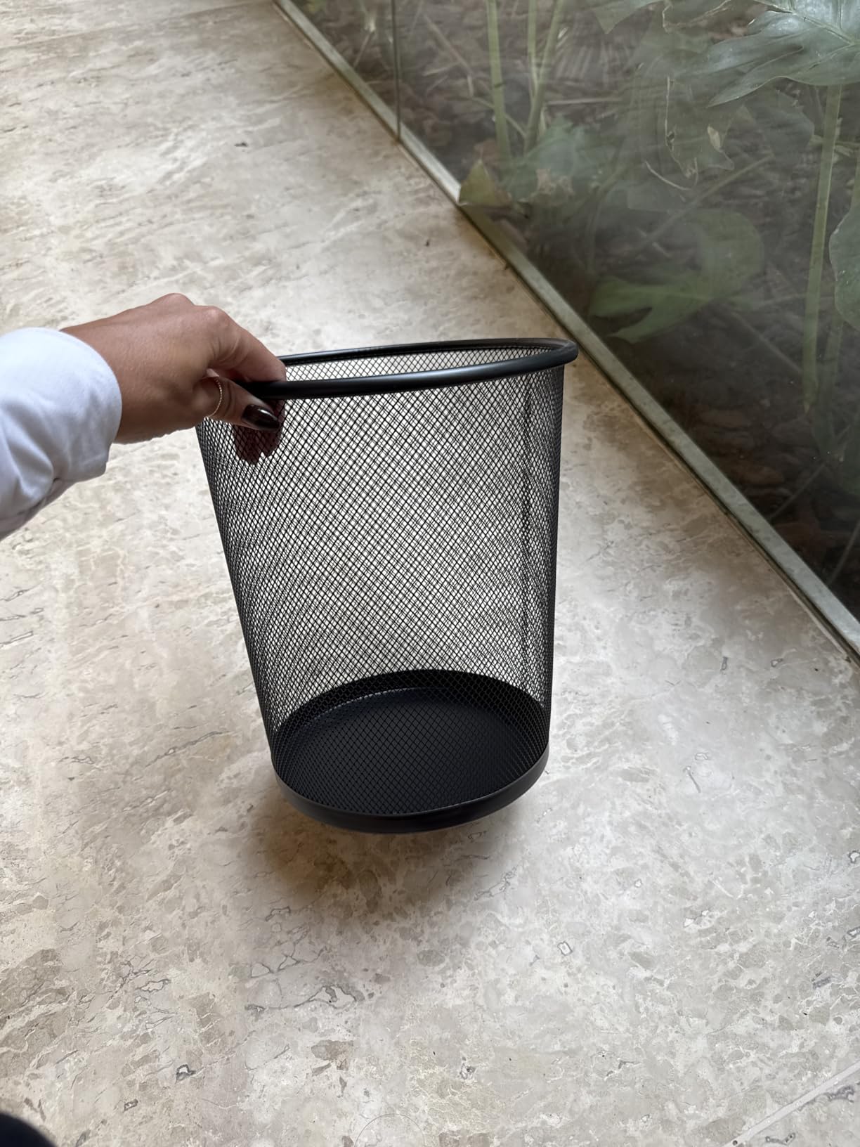 Mão de uma pessoa segurando a lixeira aramada preta sobre piso de mármore