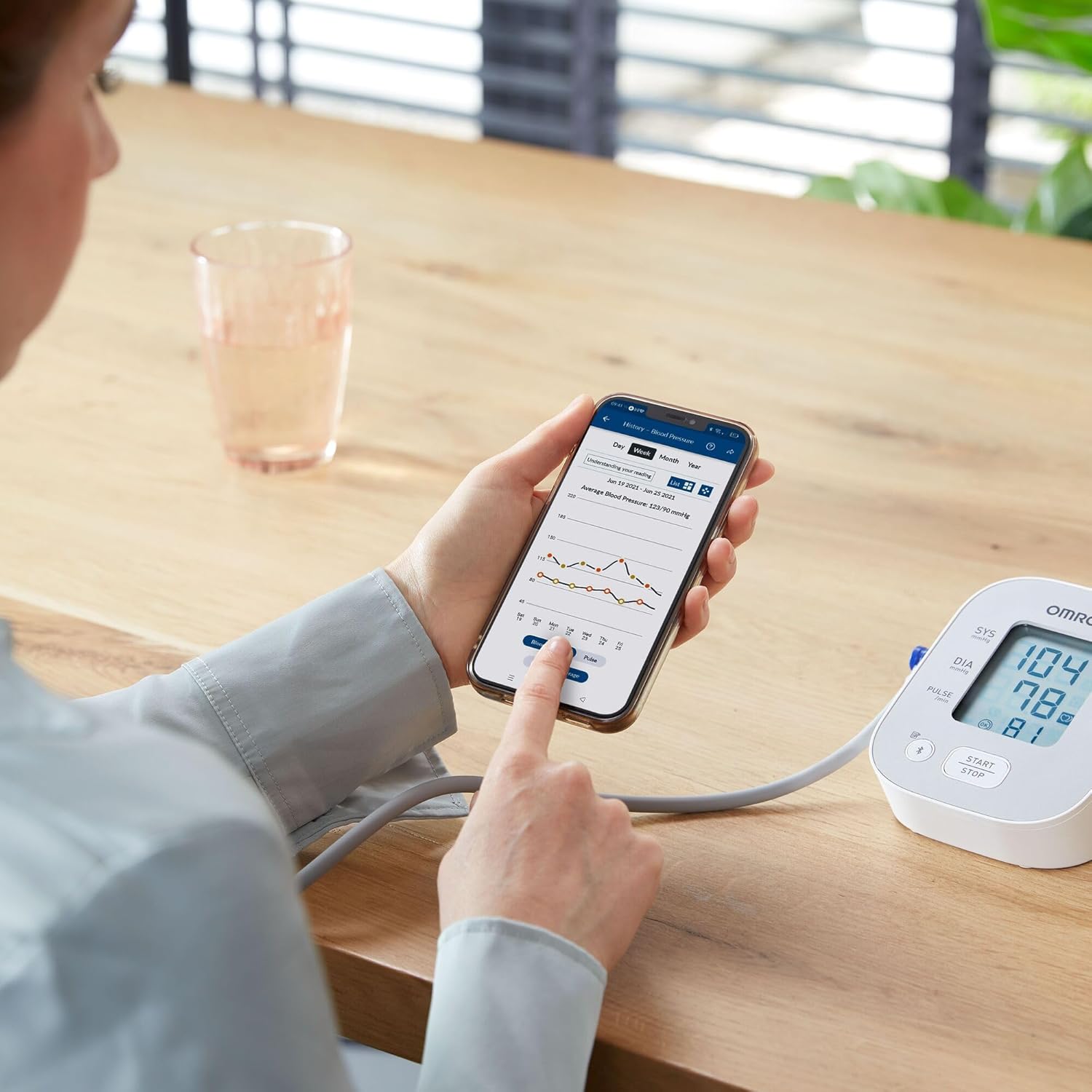 A user interacting with the OMRON connect app on a smartphone, displaying blood pressure trends and data, with the monitor visible in the background.
