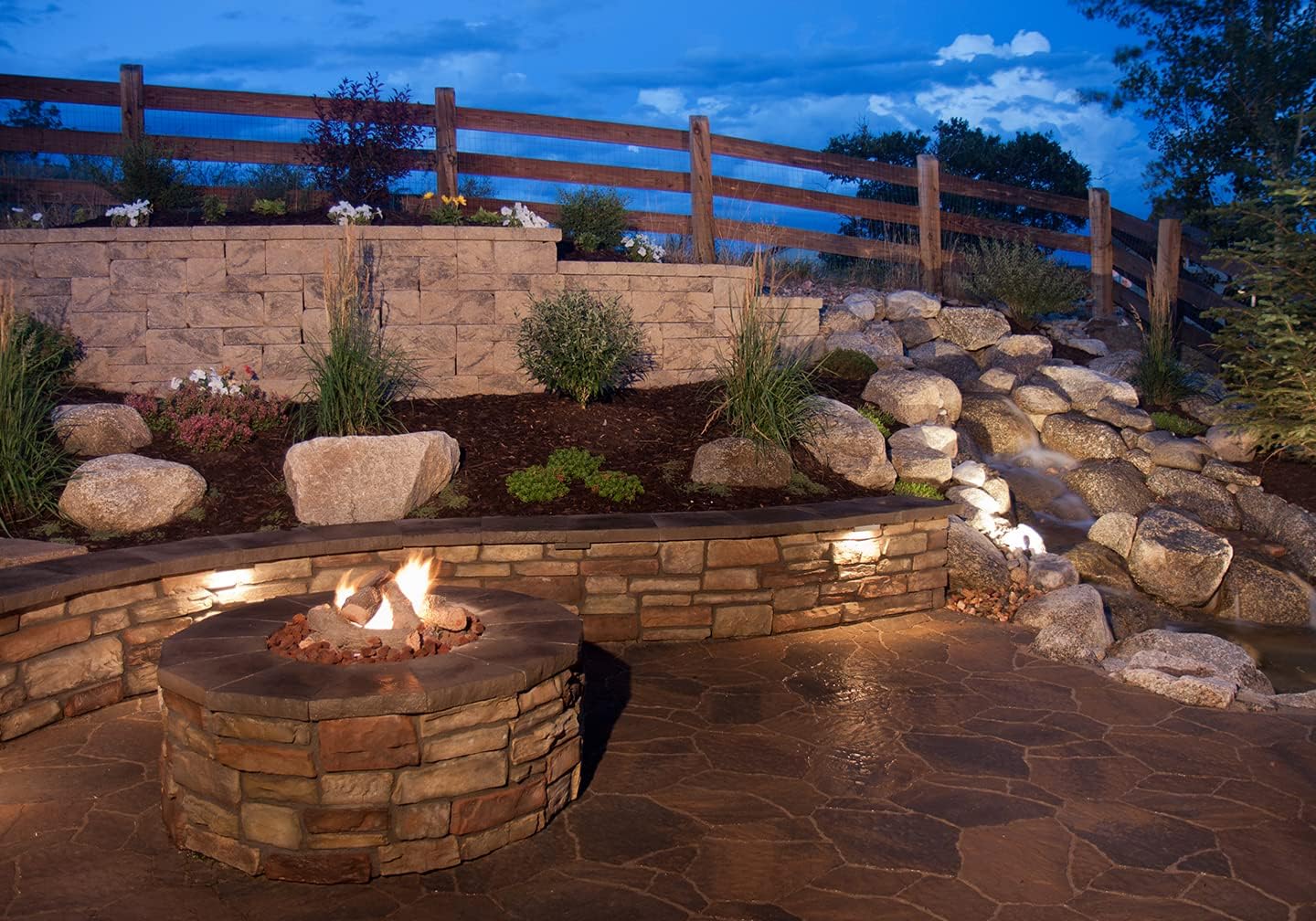 Outdoor patio scene at dusk with GKOLED hardscape lights illuminating a retaining wall and steps around a fire pit.