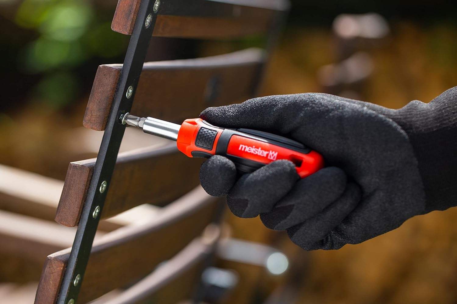 A gloved hand using the Meister screwdriver handle with a bit to tighten a screw on a wooden slat.