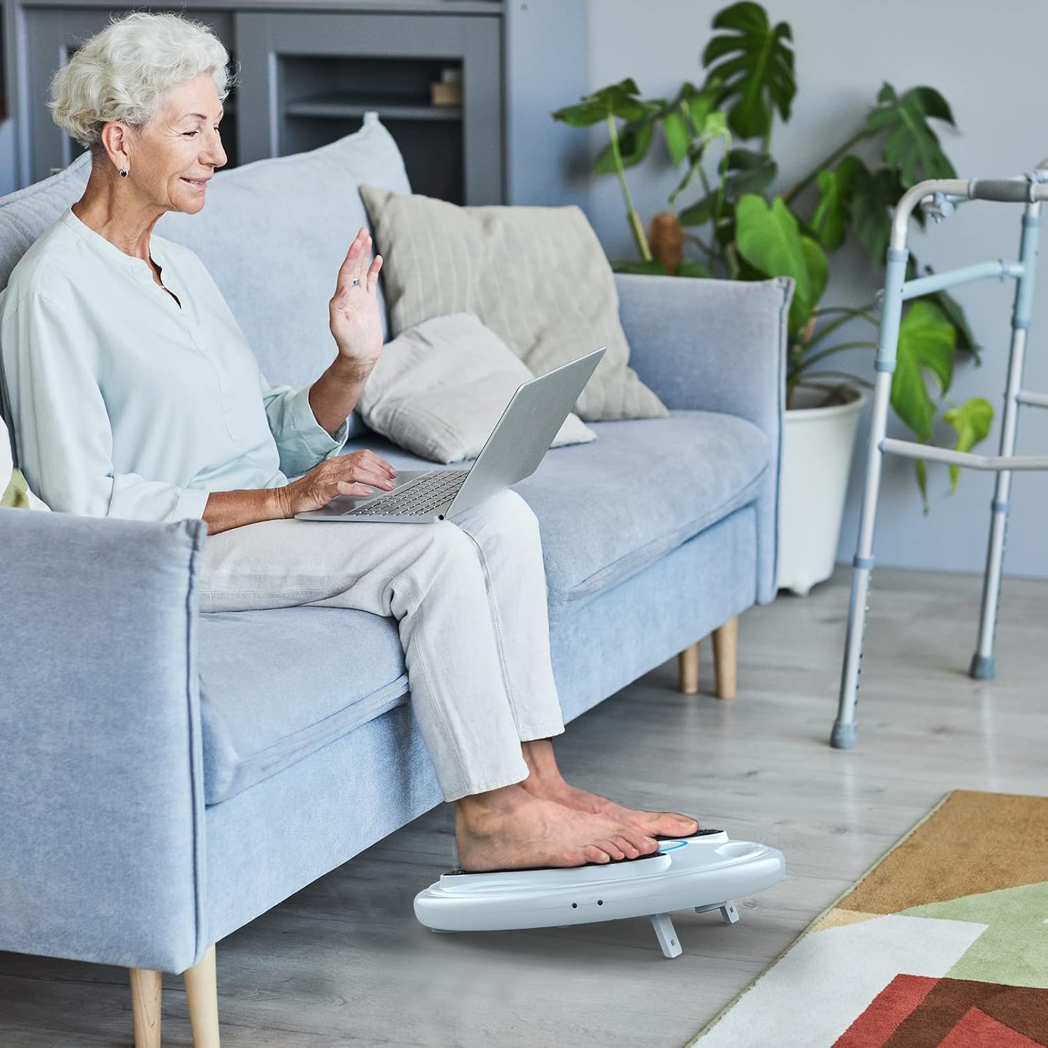 An elderly woman sitting on a couch, using the OSITO foot circulation device with her feet placed on the pads.