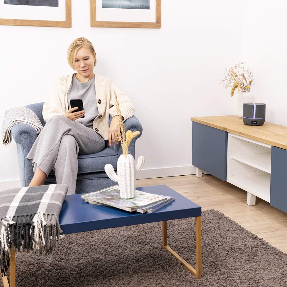 A person relaxing on a chair, looking at their smartphone, with the Cecotec PureAroma Diffuser 550 visible in the background.